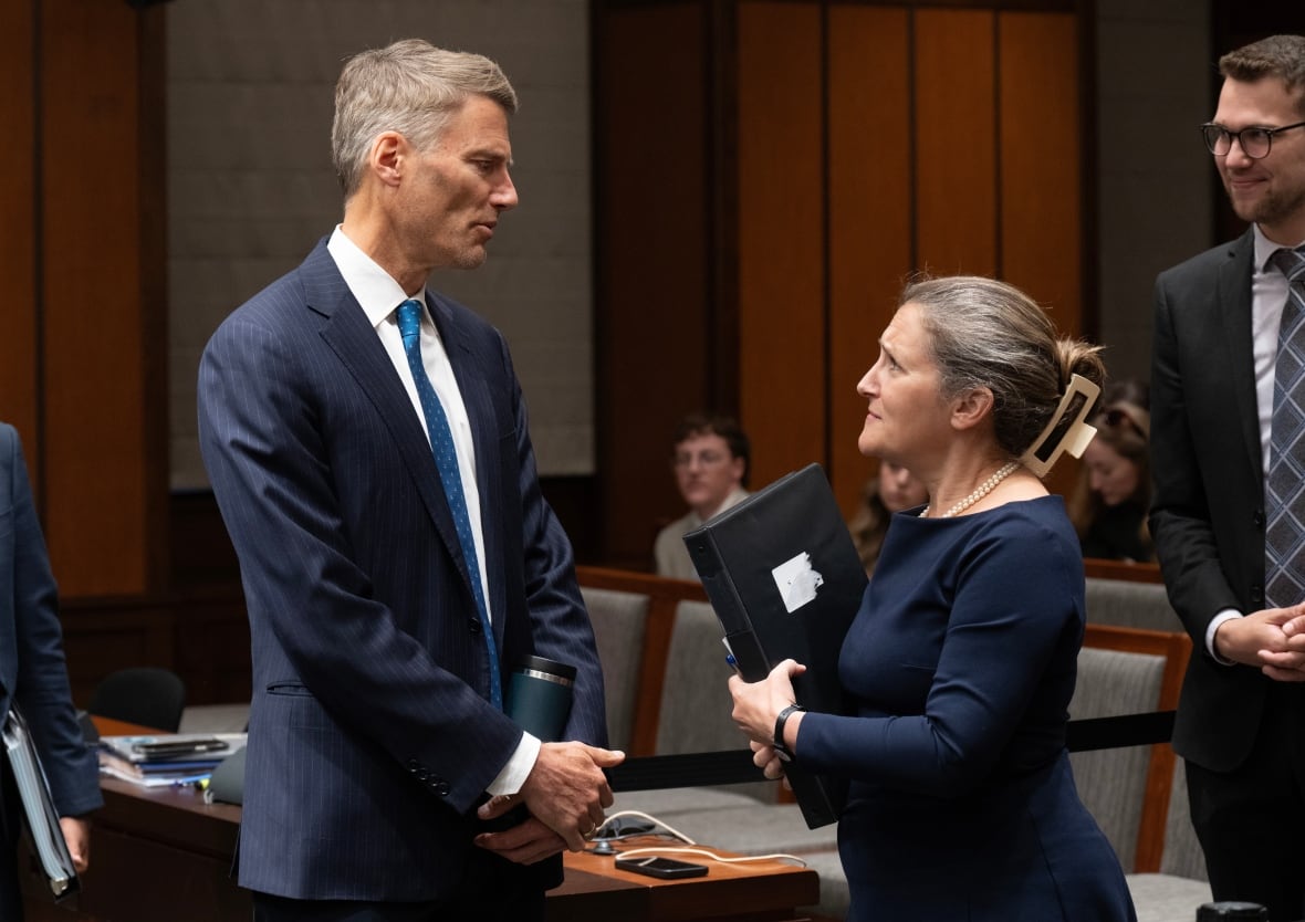 A tall man in a suit speaks with a shorter woman in a black dress inside a meeting room.