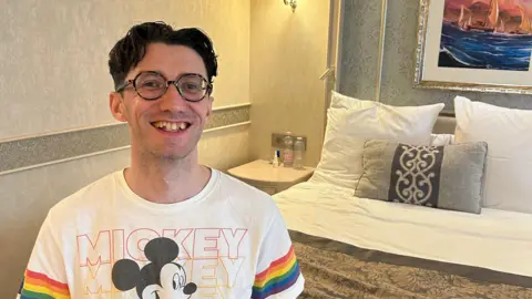 Joey Knock A man wearing round tortoise shell glasses smiles at the camera, perched on the end of a hotel bed. He is wearing a white t-shirt adorned with rainbows on each arm and a picture of Mickey Mouse on the front with the word "Mickey" repeated in different colours in the background.