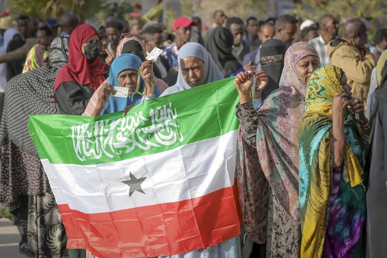 A group of women in hijabs hold up a green, white and red striped flag that has a black star in the middle and the Shahada in white calligraphic script