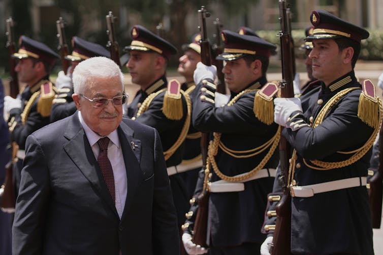 An older Palestinian man in a suit walks by a row of soldiers standing at the ready