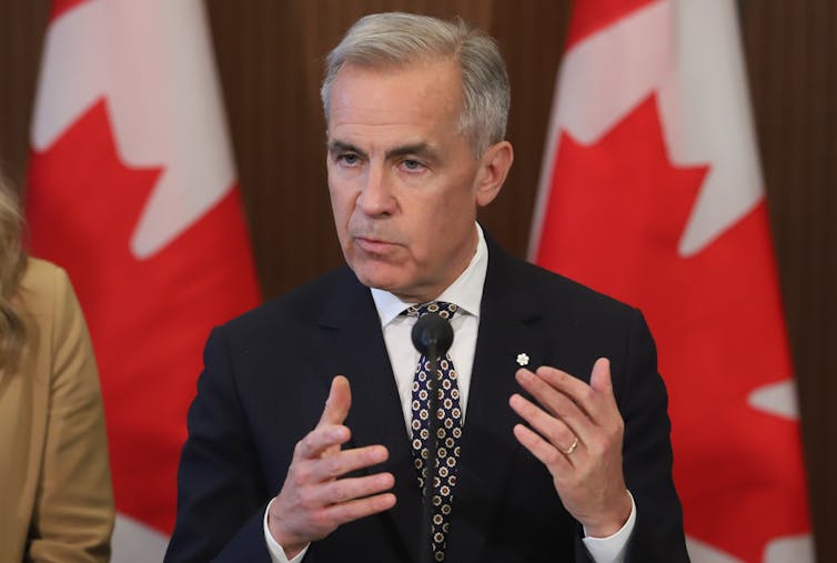 A white man in a suit speaks from behind a podium while Canadian flags are seen standing behind him