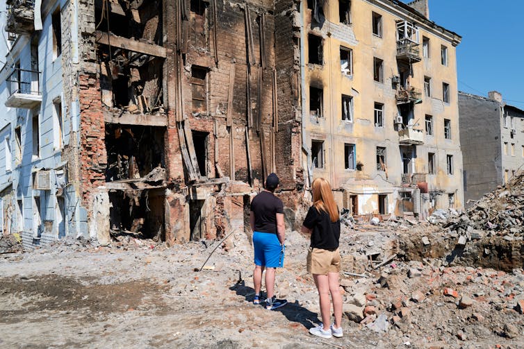 Two people standing on rubble next to bombed-out multistory buildings.