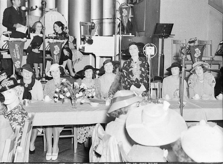 A large group of 1930s women in dresses and hats sitting at long tables in a hall