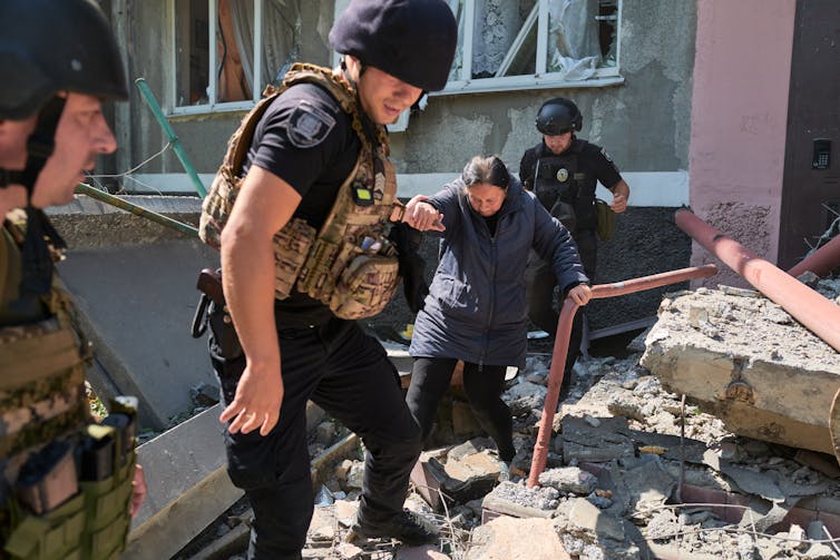Police dressed in combat gear help an old woman across rubble left after a bombing.