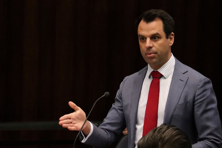 A man in a suit speaks in front of a black background.