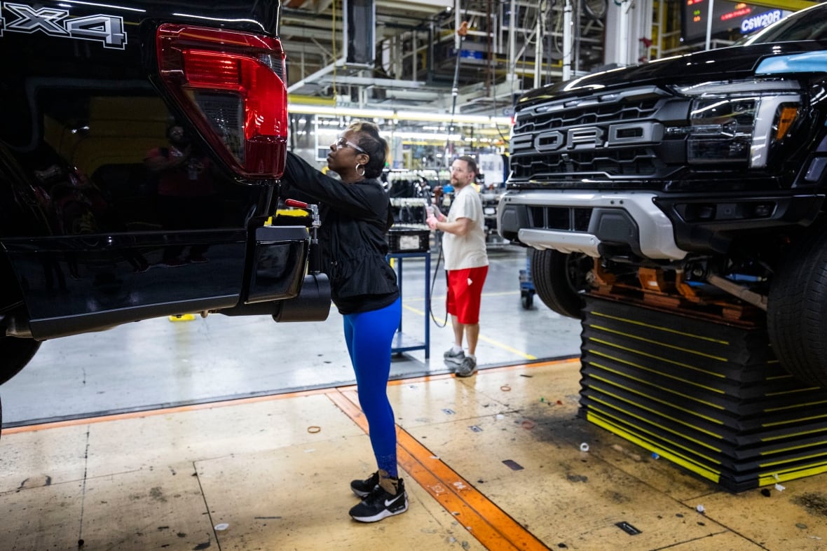 A woman in blue leggings and a hoodie wears protective eyewear as she works on a pickup truck on a vehicle assembly line while a man in red shorts looks on. 