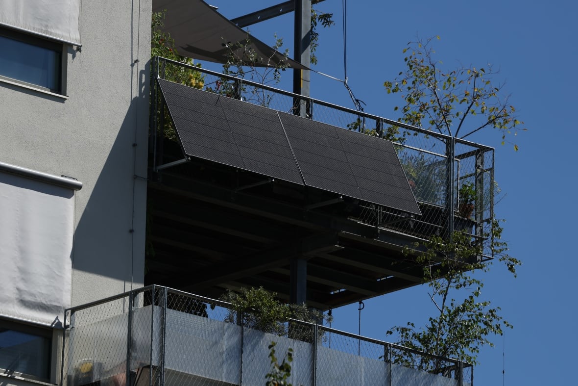 A solar panel hangs of the side of a balcony on an an upper floor of a grey, concrete apartment building. 