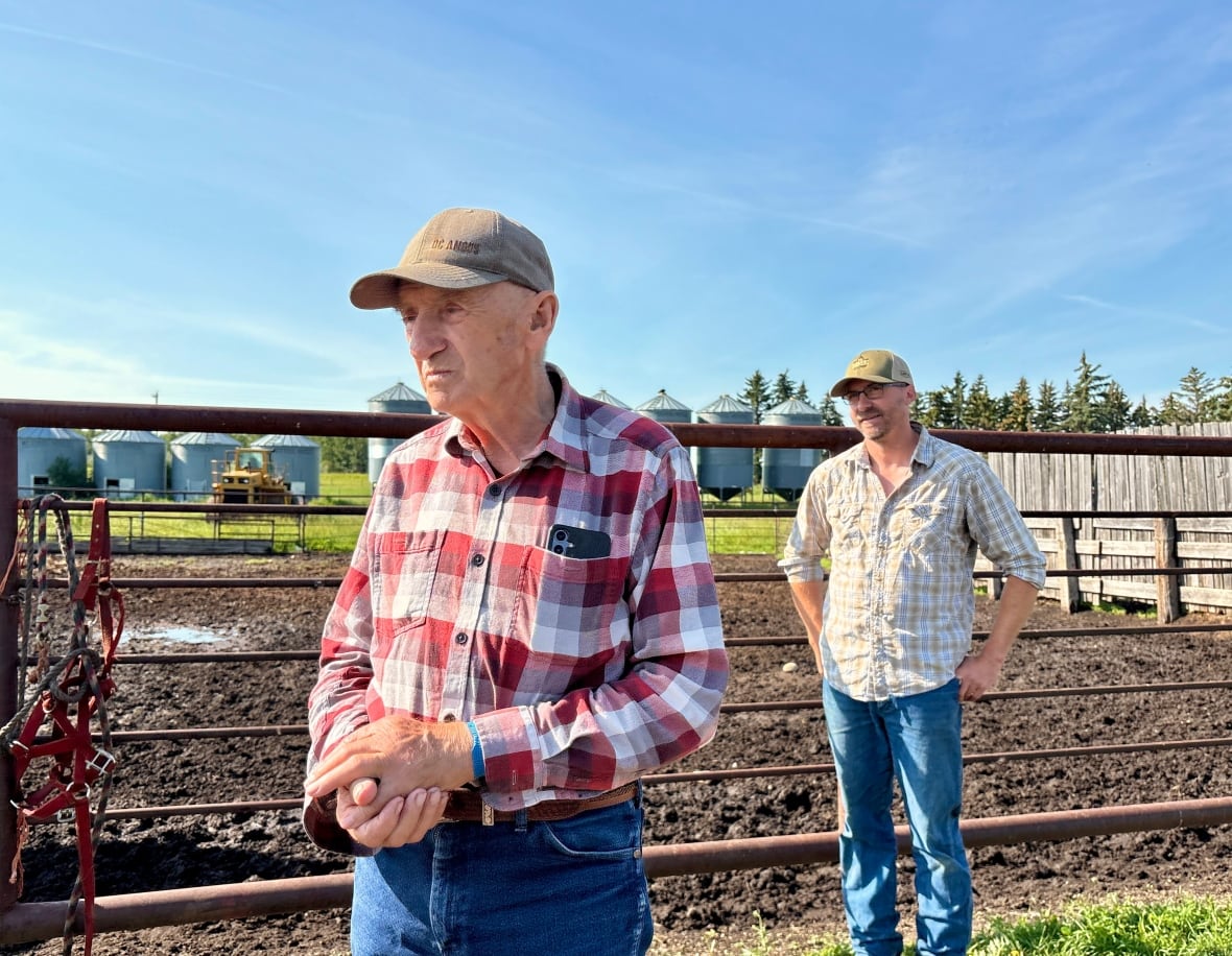 Two men stand in the dirt.  They both wear plaid shirts and jeans and ballcaps. 