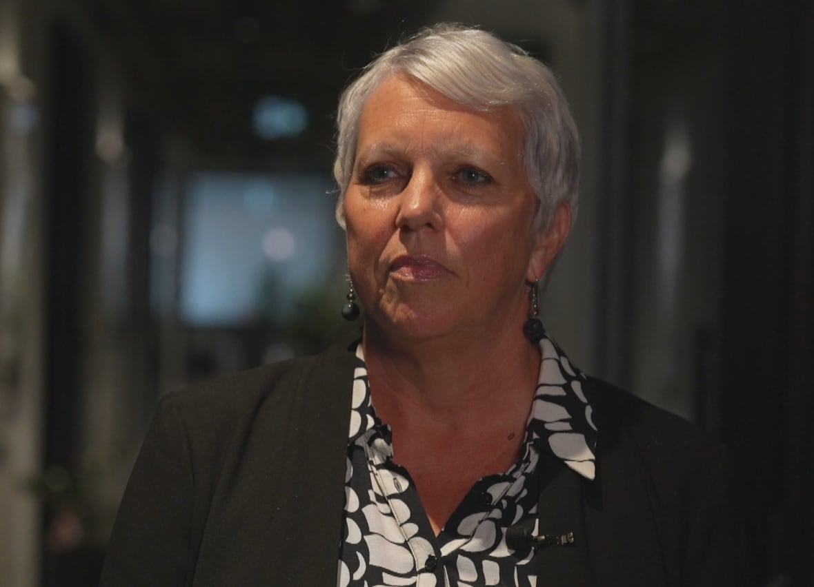 A woman with short silver hair, wearing a black and white patterned top and dark blazer, looks off-camera to the left.