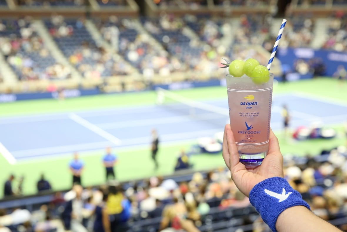 A hand holds a  cocktail in front of a tennis match