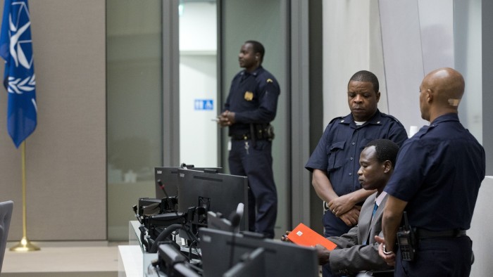 Dominic Ongwen sits at a desk in the International Criminal Court flanked by three security officers, holding a red folder.
