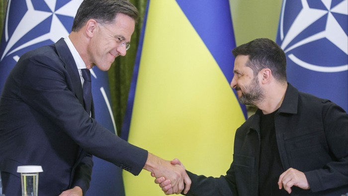 Mark Rutte and Volodymyr Zelenskyy shake hands and smile during a press conference, with Nato and Ukrainian flags behind them