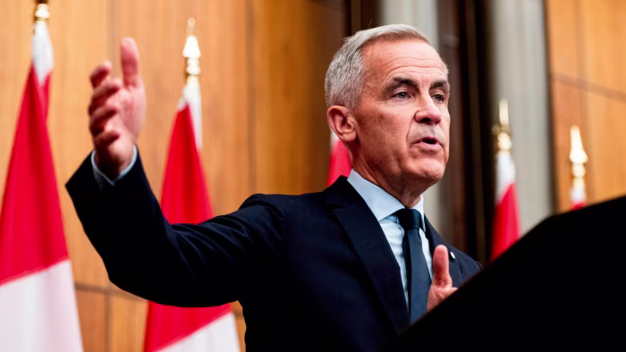 Prime Minister Mark Carney gestures while speaking at a news conference, with Canadian flags in the background