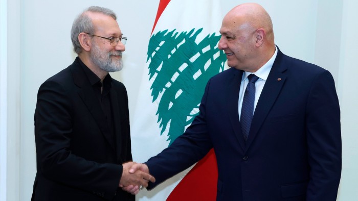 Lebanese President Joseph Aoun, right, shakes hands with Ali Larijani in front of a Lebanese flag.