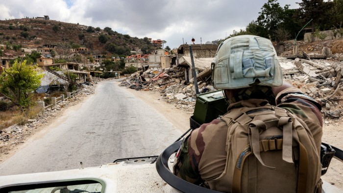 A UN peacekeeper in uniform sits atop an armored vehicle, patrolling a road lined with heavily damaged buildings