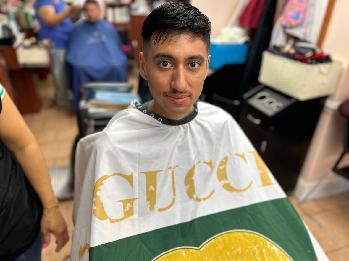 Jelver, a Hispanic man in his twenties, sits in a barber's chair wearing a green, white and gold cape that says Gucci on it. 