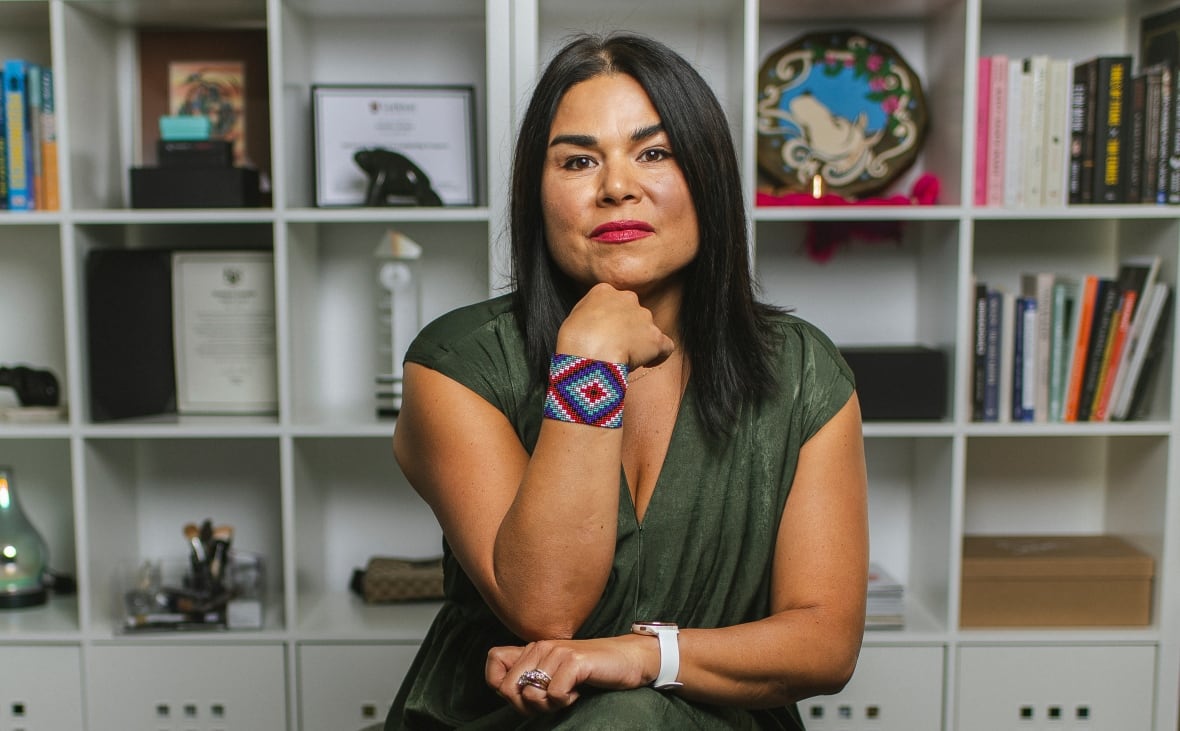 Jenn is seated in front of a wall of shelving filled with books and other items. 