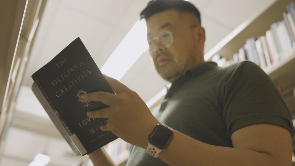 A man in glasses and a short-sleeved polo shirt peers down into an open book in a library.