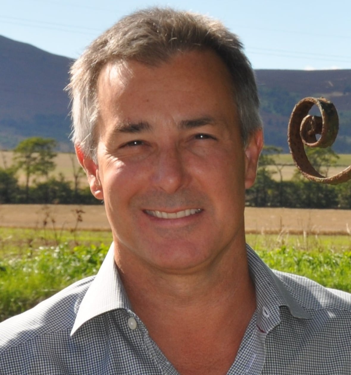 A smiling man in a fine checked shirt smile sat the camera in an outdoor photo