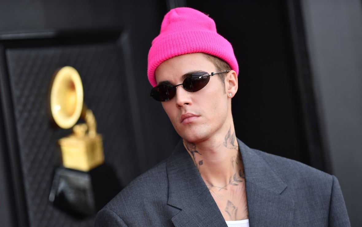 A man in a pink toque and sunglasses with neck and chest tattoos, wearing a grey suit jacket.