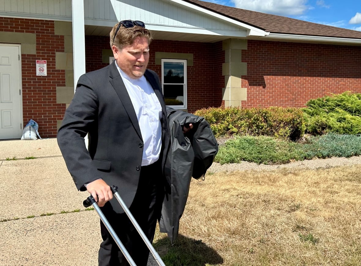 A man with brown hair and a black suit looks down as he wheels a suitcase in front of the camera.