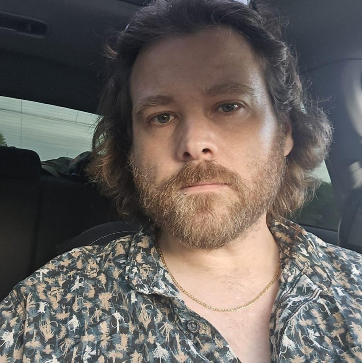 A man with a brown beard and shoulder length hair wearing a patterned shirt and a chain necklace looks at the camera.
