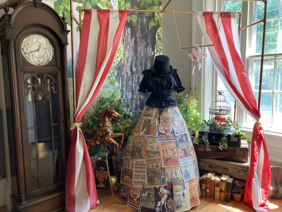A dress mannequin with magazines making up the skirt. Red and white striped curtains are tied up around the mannequin. Beside the curtain is a brown, wooden grandfather clock.