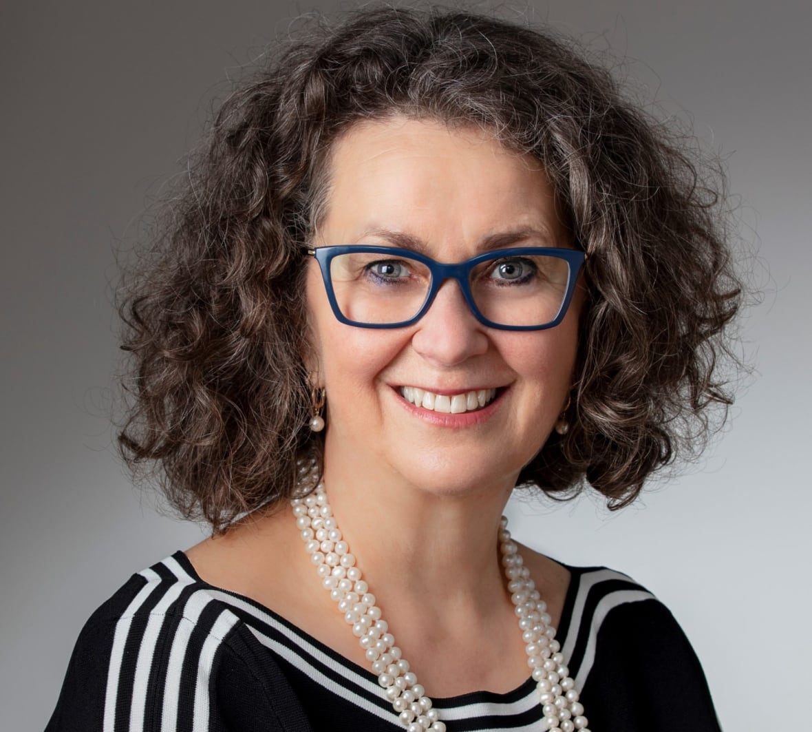 A woman in a black blouse, white pearl necklaces and blue glasses smiles for the camera. 