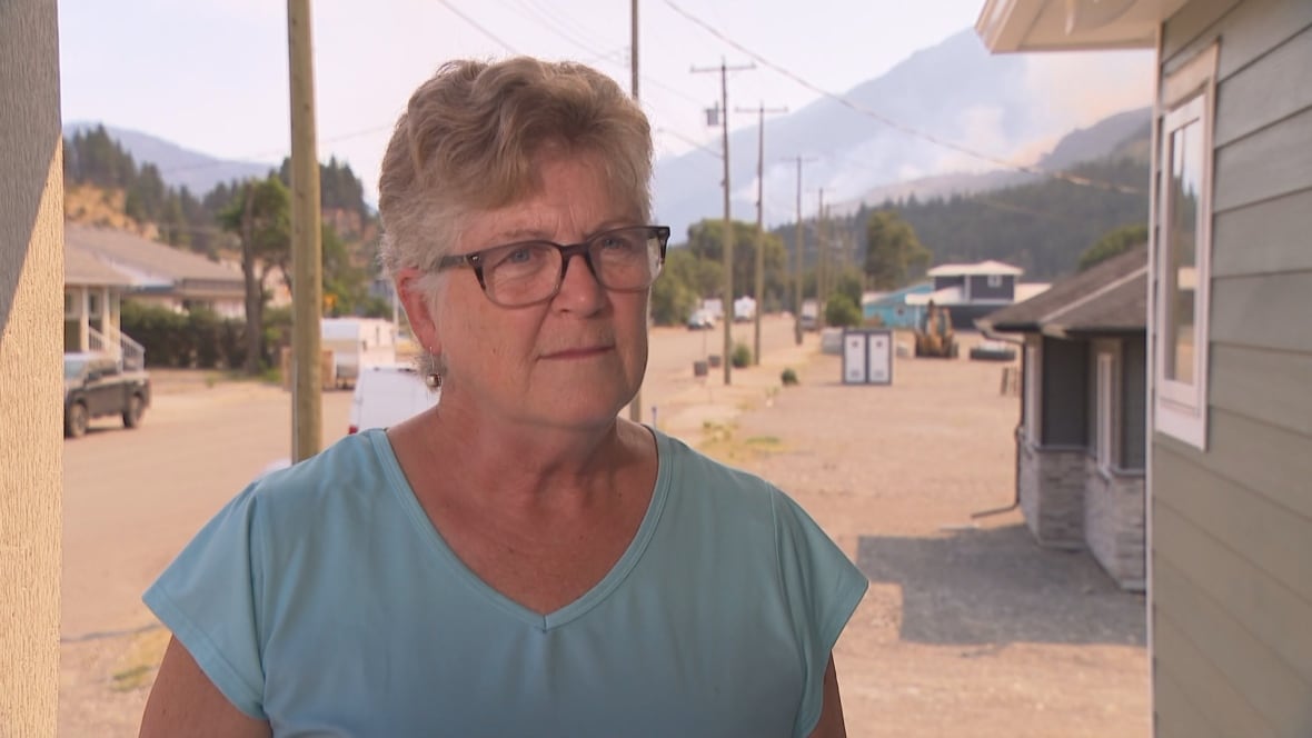 A woman with short blond hair and glasses stands outside, dressed in a blue T-shirt.