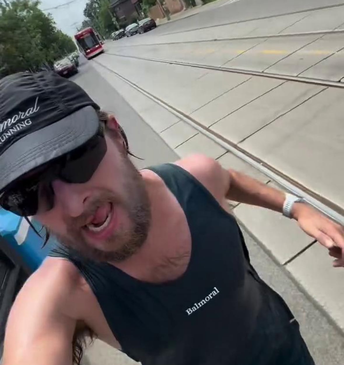 A sweating, bearded man in sunglasses films himself running selfie-style with a red streetcar on his tail.