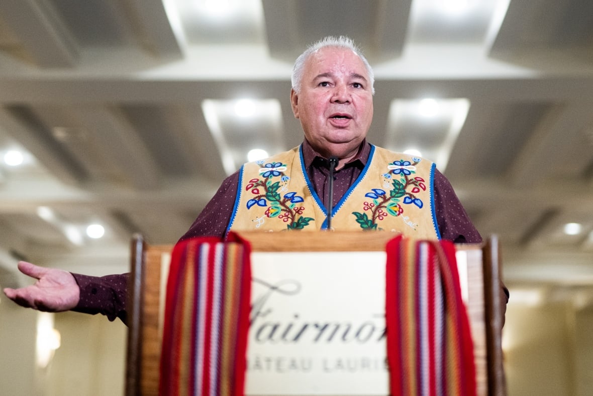 A man stands at a podium which is draped with a Métis sash.