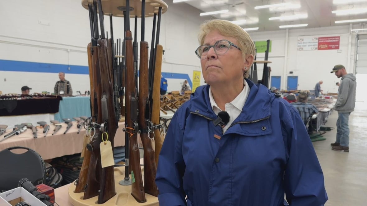 A woman wears a blue coat in front of a row of firearms.