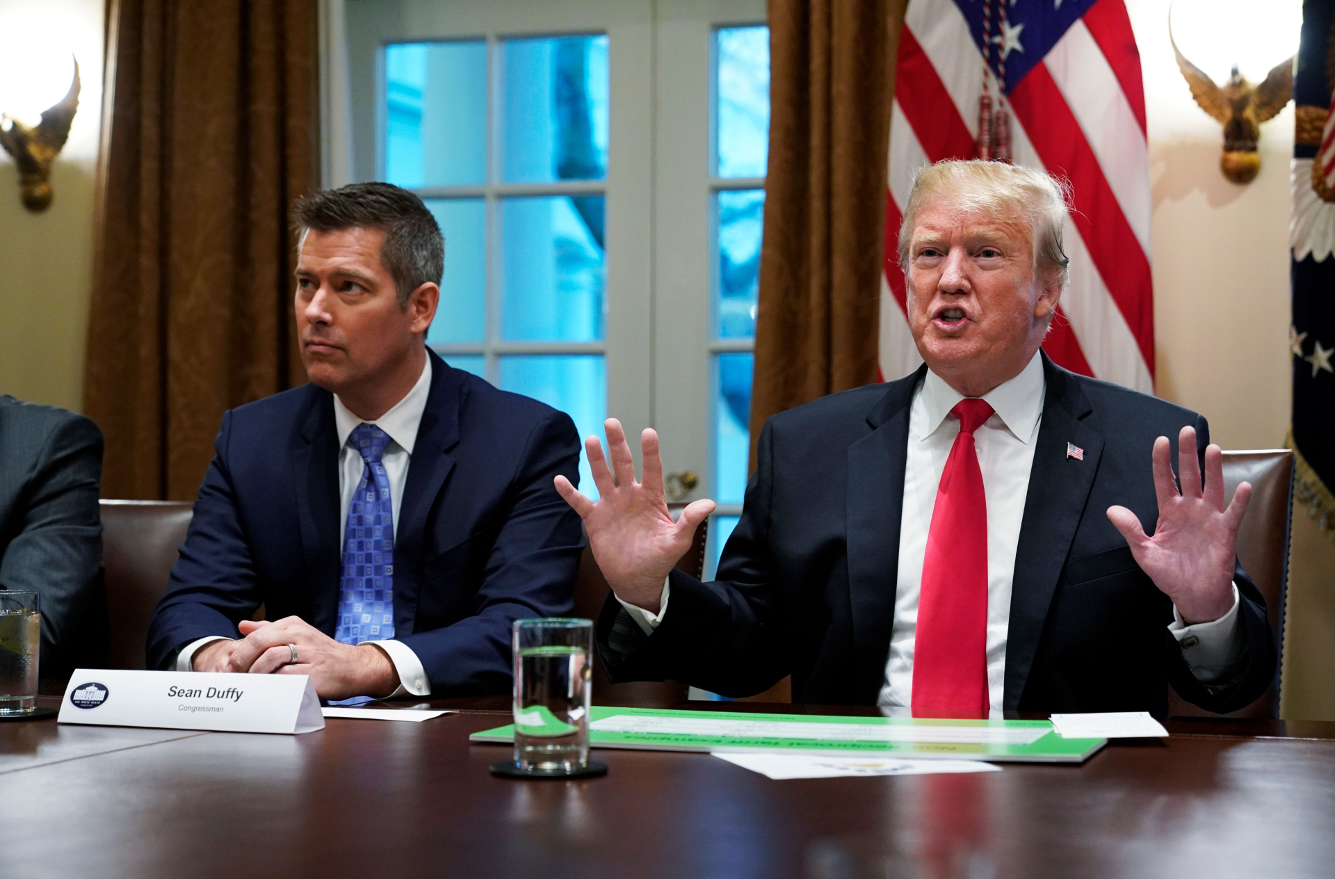 Two men wearing suits sit next to each other behind a brown table