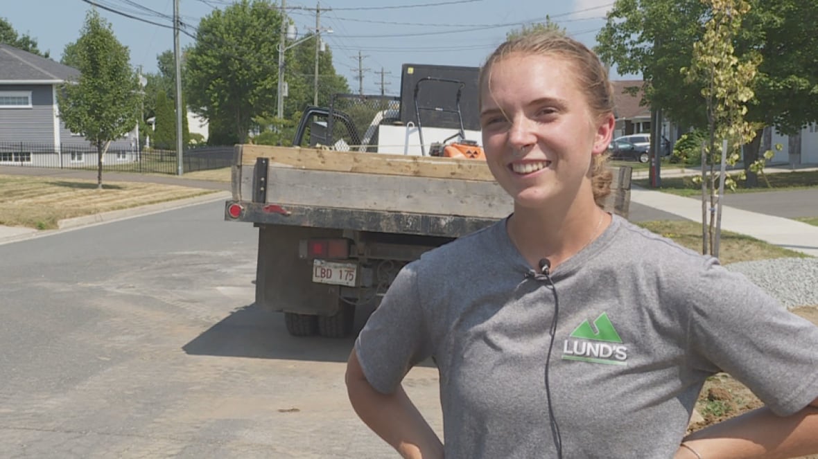 A young woman with fair hair tied back, wearing a grey T-shirt, standing in front of a work truck, on a sunny day.