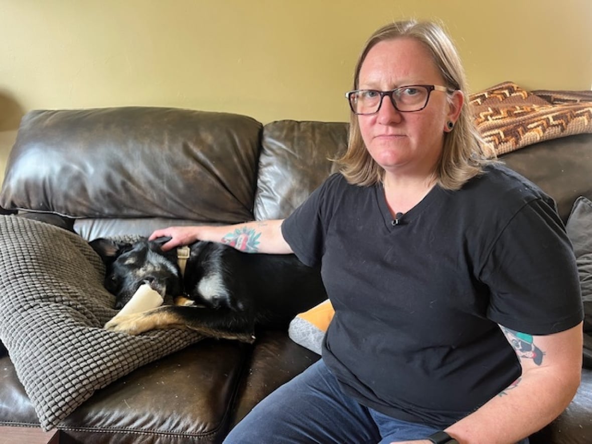 A woman wearing a black t-shirt sits on a leather couch with her arm over a black dog with beige feet and white patches in the fur near his head.