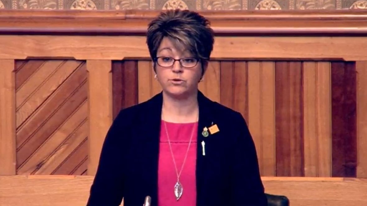 A woman with short, dark hair and glasses, wearing a dark blazer and fuchsia top, standing, speaking.