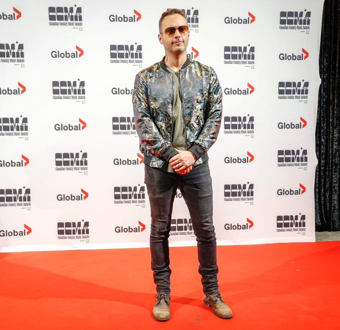 A man wearing a flashy jacket and sunglasses poses on the red carpet