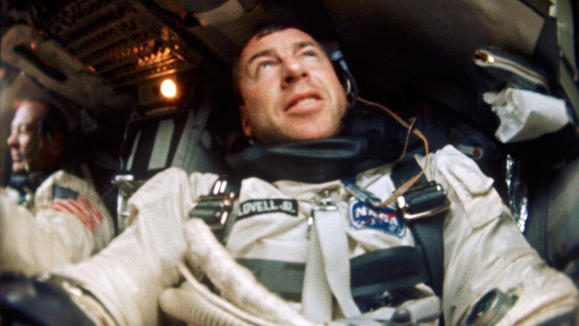 a man wearing a spacesuit looks up while seated in a cramped spacecraft