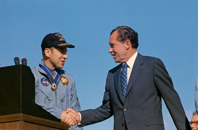 a man in a blue flight suit and ball cap shakes hands with a man in a business suit outside under a clear blue sky