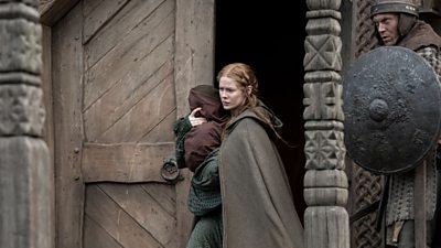 Edith Swan-neck (Emily Beecham) is walking out of a castle holding a small child. There is a guard behind her with a shield.