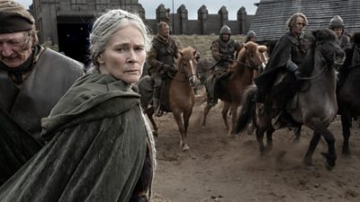 Gytha (Clare Holman) is featured on the left side of the image looking back over her shoulder. In the background, men ride horses past her.