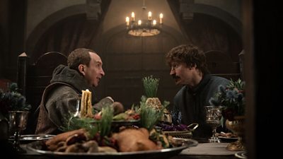 William Duke of Normandy (Nikolaj Coster Waldau) and King Edward (Eddie Marsan) sit across from one another at a table with food on