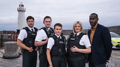 Five people stand together outdoors on a stone pier, with a white lighthouse and a police car in the background. Four of them wear police uniforms with white shirts, black ties, and black protective vests, while the fifth person, on the far right, wears a dark blazer over a mustard shirt.