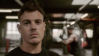 A man with short hair wearing a black sweater, standing in a boxing gym, with a focused expression looking at the camera. In the background, another man is practicing boxing on a heavy bag.