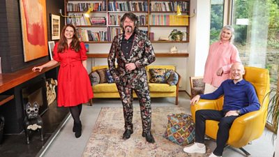 Four people pose in a modern living room with a large window and a wall of bookshelves. On the left, a woman in a bright red dress stands by a wooden shelf. In the centre, a man wearing a patterned suit with a black shirt stands with his hands in his pockets, smiling. To the right, a woman in a pale pink shirt leans on a chair, and beside her, a man in a blue sweater and white trainers sits in a mustard yellow armchair. The room features a patterned rug, colourful cushions on a yellow sofa, and contemporary artwork on the wall.