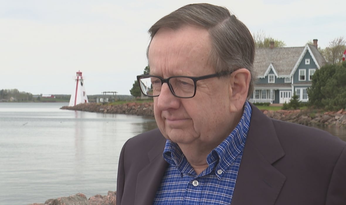 Man stands near water where a lighthouse can be seen in the background.
