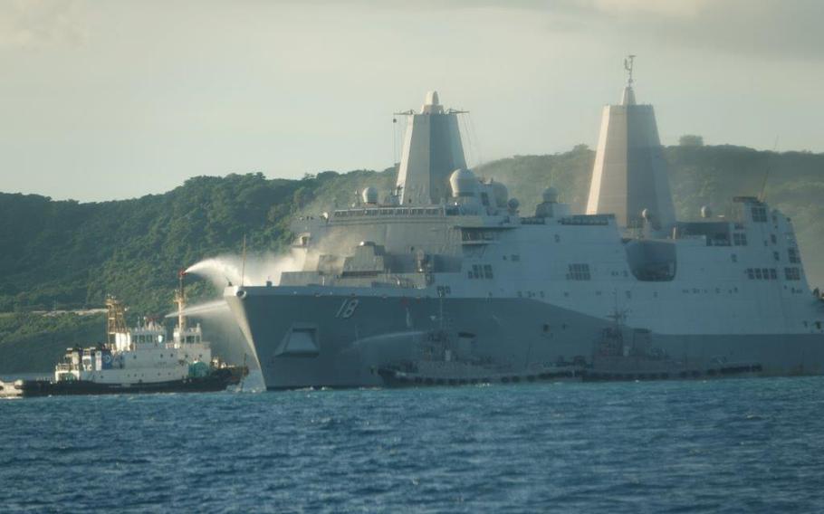 The USS New Orleans amphibious transport dock receives water sprays from a Japan Self-Defense Force vessel and two tugboats near White Beach Naval Facility in Okinawa. The large gray warship with hull number 18 is anchored in blue waters with forested hills visible in the background. 