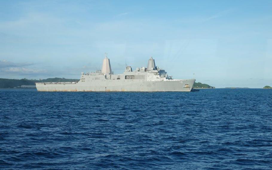 The USS New Orleans amphibious transport dock operates in deep blue waters off the coast of White Beach, Okinawa. The San Antonio-class warship displays its characteristic twin superstructure design and light gray hull against a clear blue sky. Forested islands are visible in the background, showing the tropical Pacific setting near the Okinawan coast.
