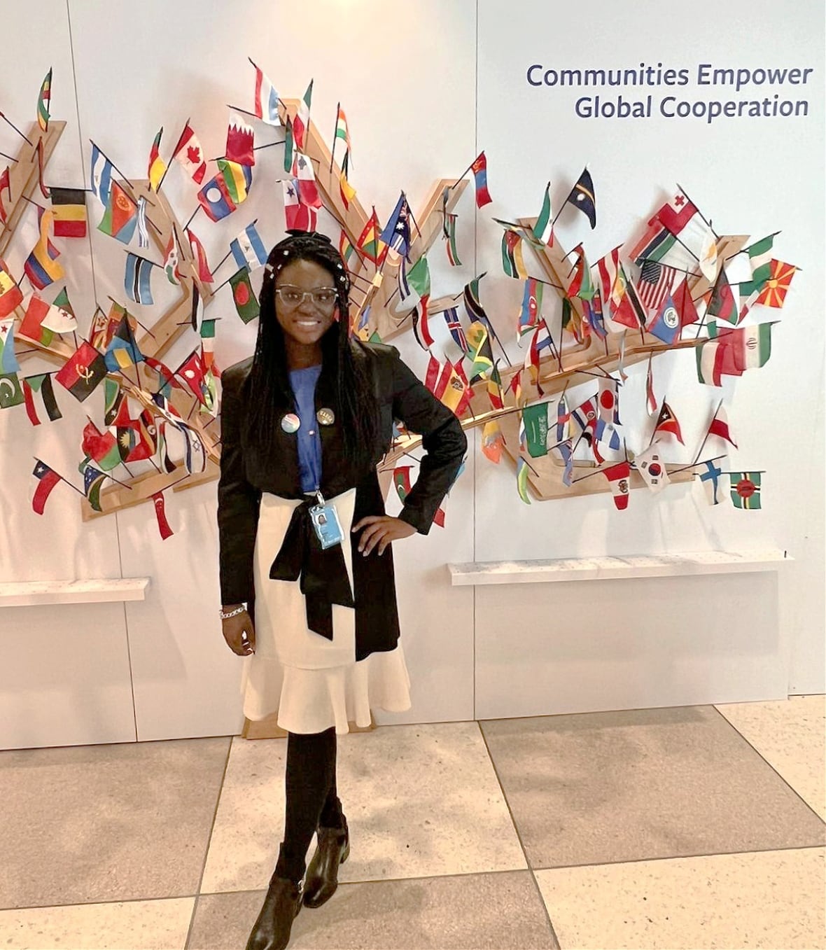 A smiling woman with long hair, wearing a black jacket and white skirt, stands before a display of national flags and the message: Communities empower global cooperation.