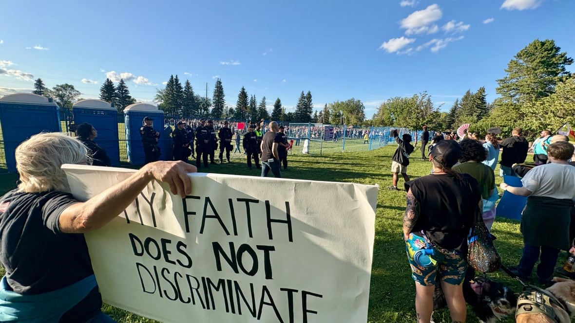 Female protestor with a sign that reads "My faith does not discriminate" stands outside of concert venue with several other protestors.